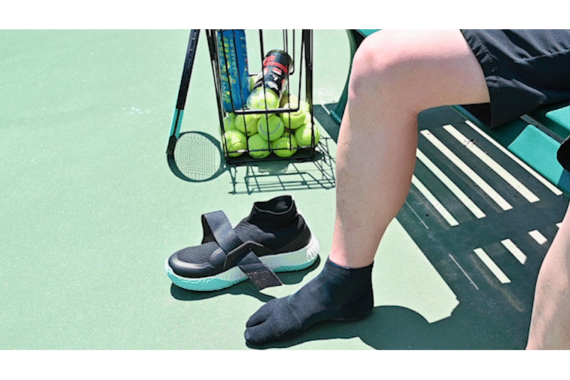 A gif of a man sitting on a bench on a tennis court putting on the black/white colorway of The One and strapping it onto his foot.
